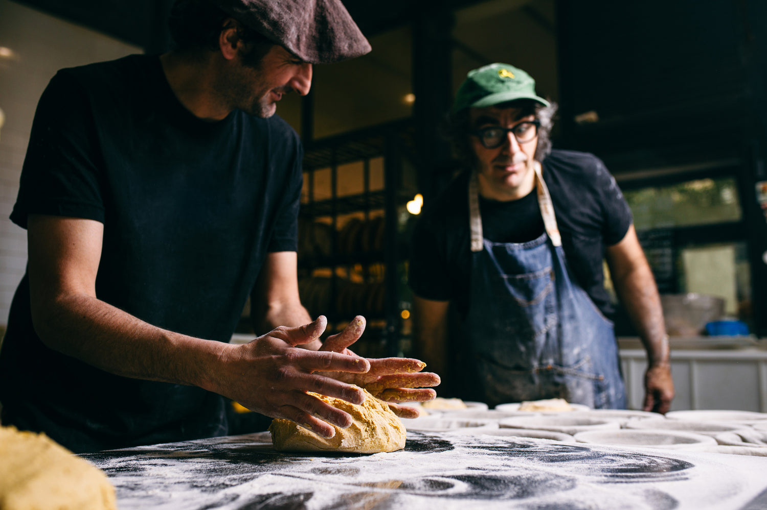 One person works dough on a flour covered table as another looks on